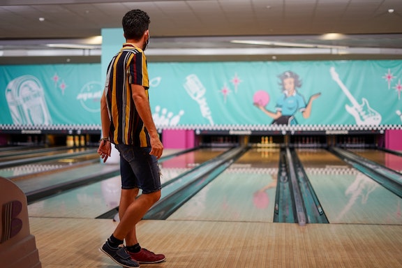 A person with short hair in a striped yellow shirt looks down the lane of a bowling alley, presumably having just bowled.